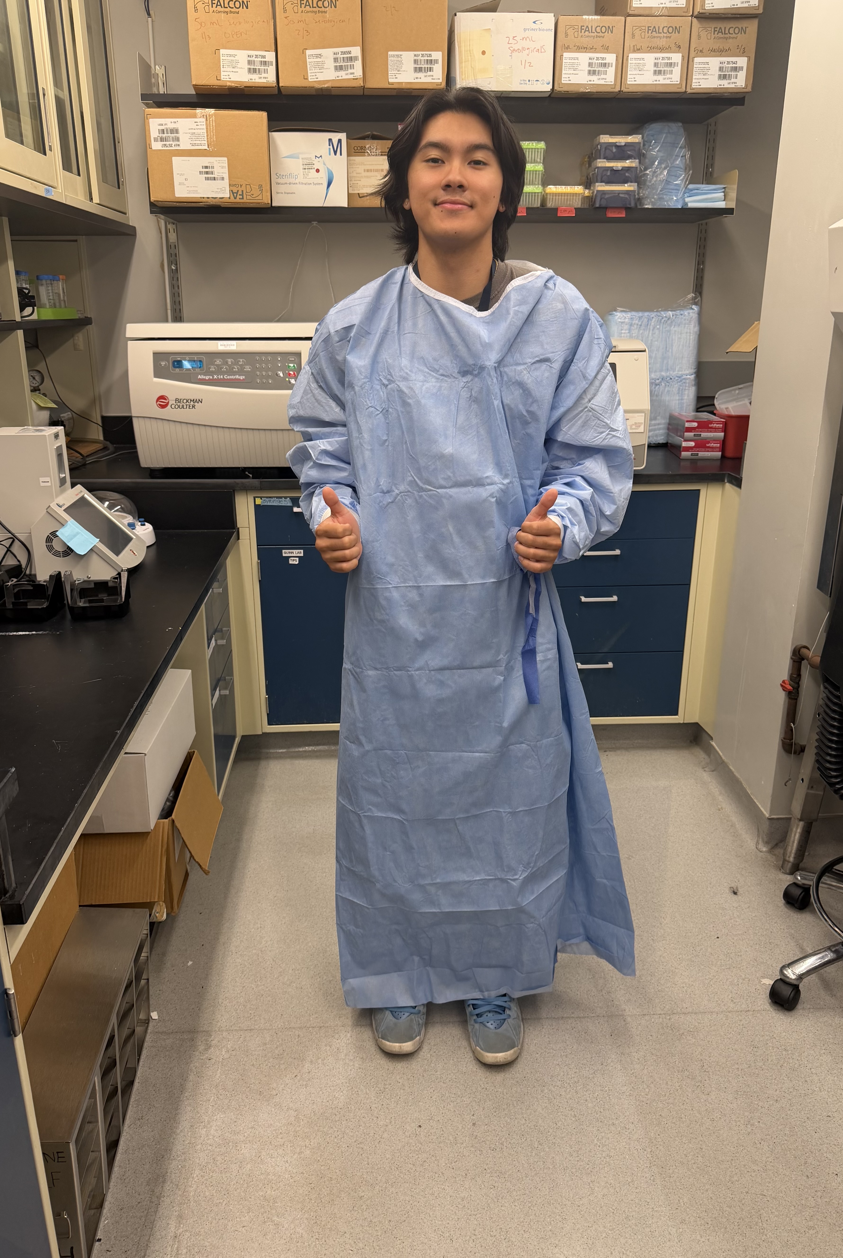 A young man wearing an oversized blue medical gown smiles and gives two thumbs-up inside a laboratory filled with equipment, supply boxes, and shelving.
