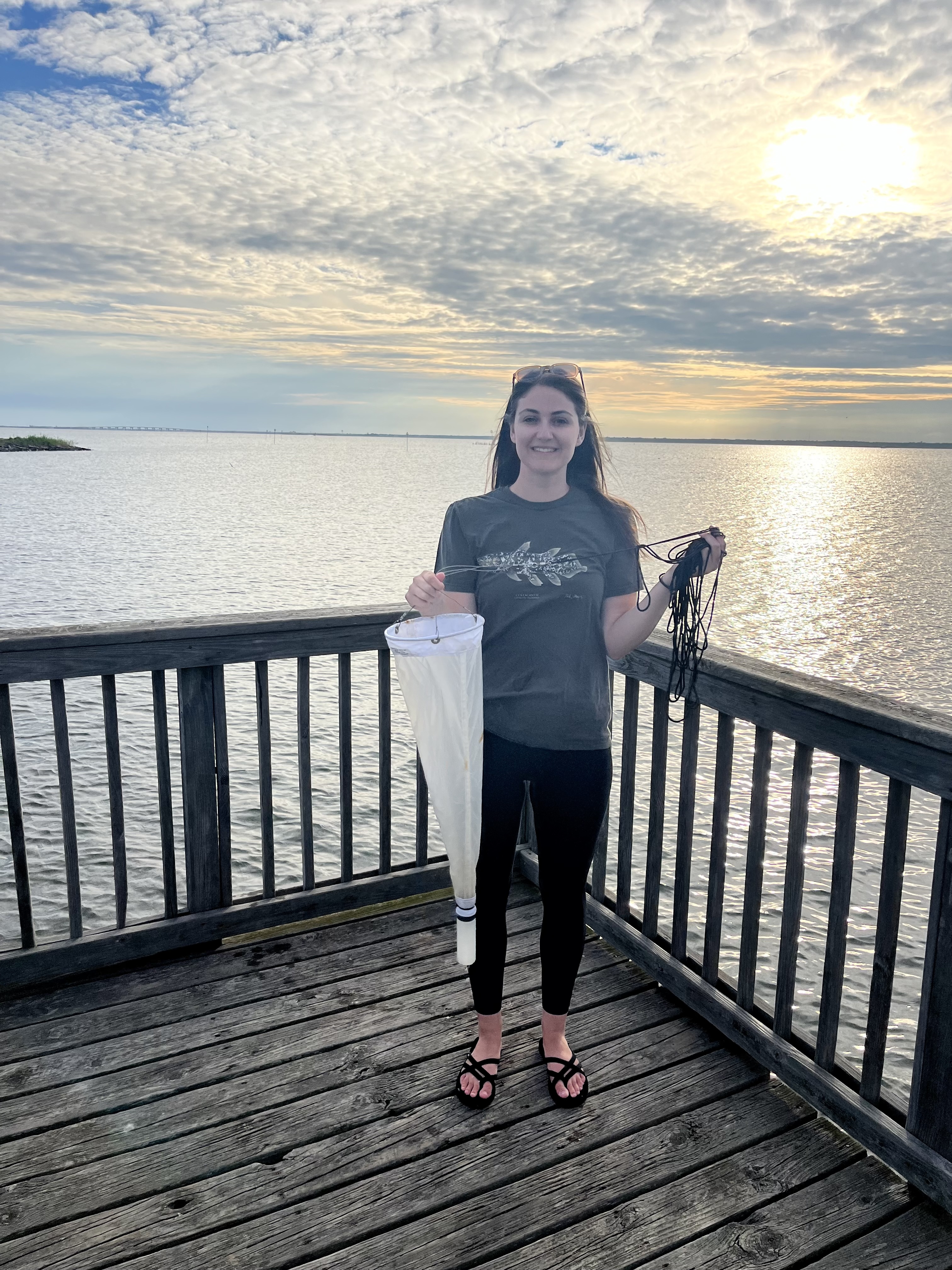 A woman standing on a wooden pier near a large body of water during sunset, smiling while holding a white conical plankton net in one hand and black cord in the other under a bright, cloudy sky.
