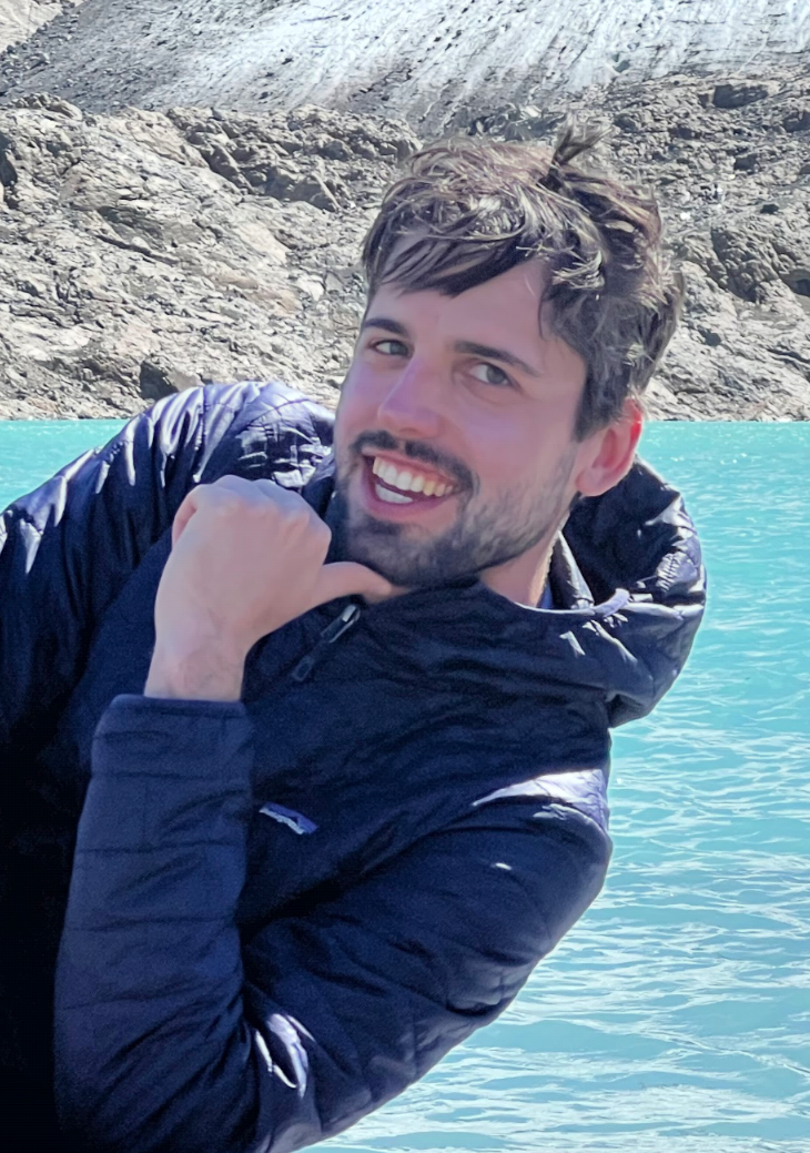 A man with brown hair and beard in a blue jacket poses smiling in front of water and rocks.