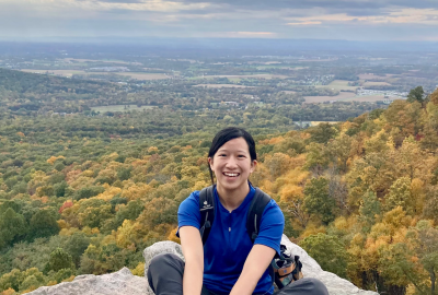 Biological sciences graduate student Stephanie Chia sits smiling at the camera on a rock overlooking a cloudy sky and a valley of trees.