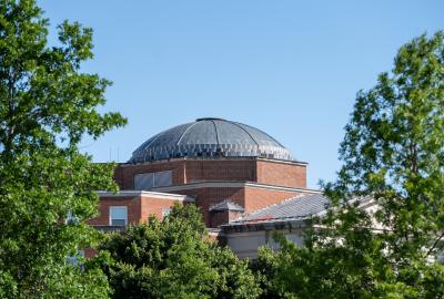 Dome of a building with red brick, surrounded by green trees, with blue sky above