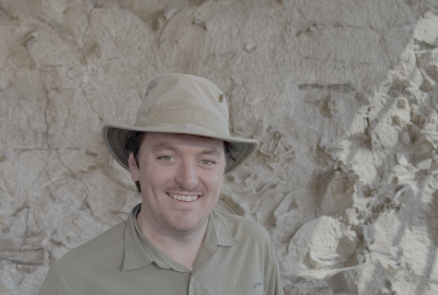 Nathan Jud in brown hat and shirt smiles while standing in front of a rock wall.