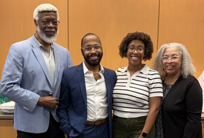 Four adults stand together smiling in front of a light wood-paneled wall. From left to right: a tall man with white hair and beard wearing a light blue blazer and black pants; a man in a navy blue suit with brown shoes; a woman wearing glasses, a white striped short-sleeve sweater, and olive green pants; and a woman with curly gray hair wearing glasses, a black cardigan, and a patterned black-and-white dress. 