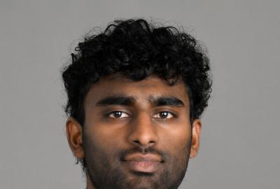 Headshot of a man wearing a white dress shirt, light blue tie and royal blue sweater in front of a gray backdrop.