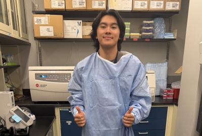 A young man wearing an oversized blue medical gown smiles and gives two thumbs-up inside a laboratory filled with equipment, supply boxes, and shelving.