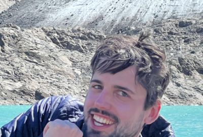 Brown haired man with beard and blue jacket smiling with his chin on his hand in front of water and rocks.