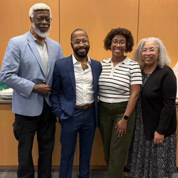 Four adults stand together smiling in front of a light wood-paneled wall. From left to right: a tall man with white hair and beard wearing a light blue blazer and black pants; a man in a navy blue suit with brown shoes; a woman wearing glasses, a white striped short-sleeve sweater, and olive green pants; and a woman with curly gray hair wearing glasses, a black cardigan, and a patterned black-and-white dress. 
