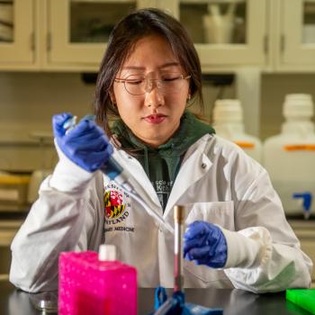 A woman in a lab coat wearing blue gloves and glasses uses a pipette to transfer liquic into a test tube.