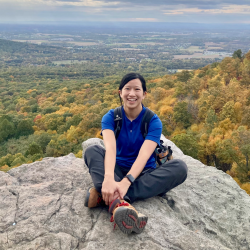 Biological sciences graduate student Stephanie Chia sits smiling at the camera on a rock overlooking a cloudy sky and a valley of trees.