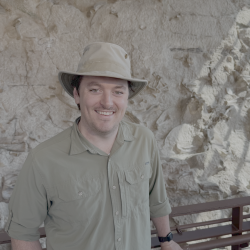 Nathan Jud in brown hat and shirt smiles while standing in front of a rock wall.