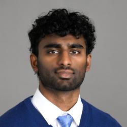 Headshot of a man wearing a white dress shirt, light blue tie and royal blue sweater in front of a gray backdrop.