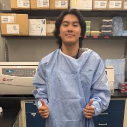A young man wearing an oversized blue medical gown smiles and gives two thumbs-up inside a laboratory filled with equipment, supply boxes, and shelving.
