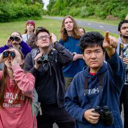 A man in a blue hoodie points as nine students behind him look through binoculars. 
